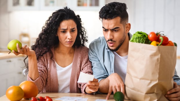 Shocking Prices. Confused Middle Eastern Spouses Checking Bills After Grocery Shopping, Young Arab Couple Sitting At Table In Kitchen, Upset Man And Woman Reading Supermarket Invoice, Closeup photo