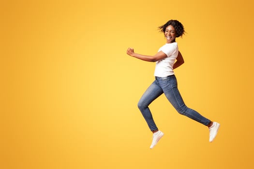 Active black lady in casual outfit running over orange studio background, full length shot. Cheerful african american young woman in jeans and white t-shirt having fun on her own photo
