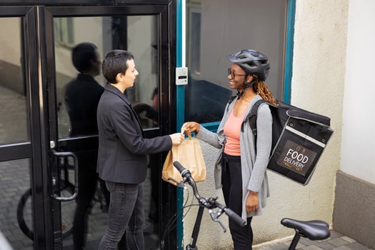 African American woman courier bringing meal order to a customer outside an apartment building. Worker ensuring express delivery service for urban takeaway shipping, delivery bike. photo