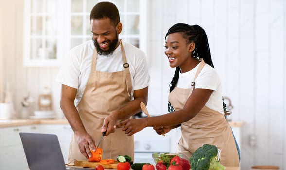 Happy African American Spouses Using Laptop Computer While Cooking In Kitchen Together, Searching Recipe In Internet, Smiling Black Couple Preparing Healthy Food For Dinner At Home, Copy Space photo