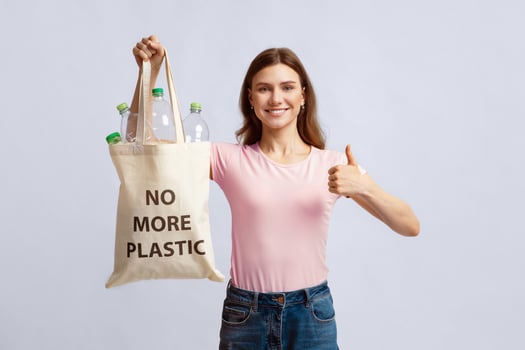 Cheerful Woman Holding Eco Bag Filled With Empty Bottles And No More Plastic Inscription, Smiling Lady Showing Thumb Up, Enjoying Zero Waste Living, Posing On Gray Studio Background With Empty Space photo