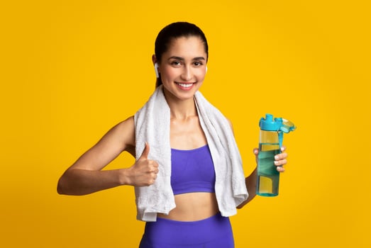 Stay Hydrated. Portrait of happy smiling female fitness instructor in wireless earphones holding bottle with fresh water and showing thumbs up sign gesture, posing isolated on orange studio background photo
