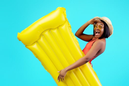 Millennial black woman in swimsuit holding yellow inflatable lilo on blue studio background. Young African American lady with pool float having tropical summer vacation or beach party photo