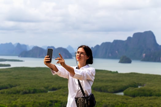Young woman taking selfie with smartphone against stunning landscape of mountains and water, capturing moment of joy and adventure in nature's beauty. Female traveler is taking selfie with smartphone photo