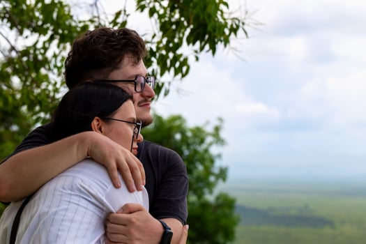 Couple embracing outdoors, surrounded by lush greenery and scenic landscape, capturing moment of affection and connection in serene environment with soft clouds above. Man gently hugs woman photo