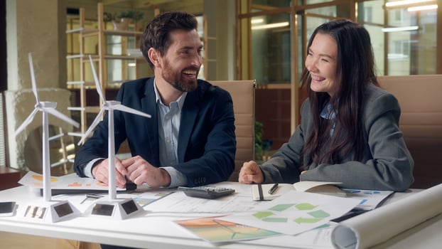 Happy joy Caucasian businessman Korean businesswoman discuss wind turbine model pointing finger laugh talk eco energy office startup sustainable development meeting cooperation innovation paper office photo