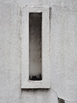 Close up of rectangular wall vent embedded in concrete wall, showing signs of weathering and dirt accumulation inside vent opening, with visible cracks and rough texture on wall surface photo