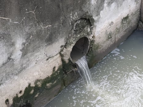 Small pipe embedded in weathered concrete wall releases water into murky water body, with moss and algae growth around pipe opening, creating natural, aged, and slightly neglected scene photo