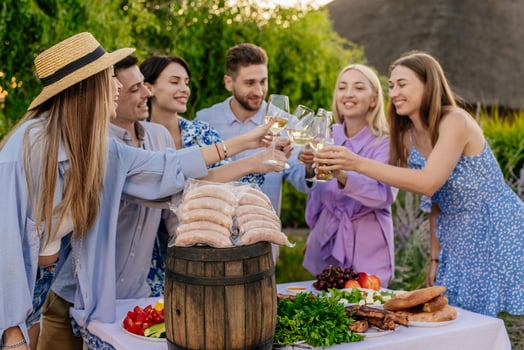 Happy friends eating, drinking beer, and raising a toast at a BBQ dinner in nature, next to sausages for the barbecue. Concept food and friendship, promoting BBQ and outdoor lifestyle photo