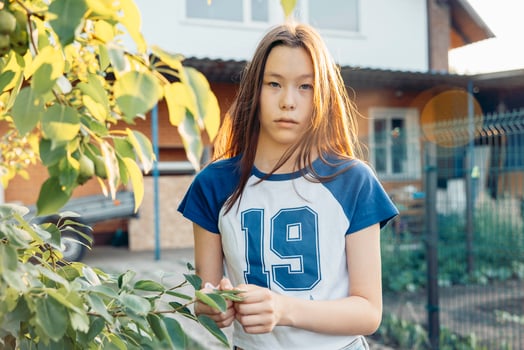Asian Metis girl holding leaves in backyard garden during sunset gazing at camera with calm expression near suburban house and green foliage concept of lifestyle brand, teen fashion, outdoor photo