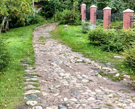 The rural cobblestone road runs along a wrought iron black metal fence with brick posts, in the middle of a park with grass and trees. photo