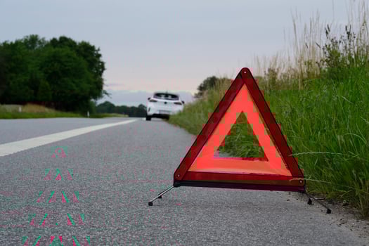 On a side of the road is a warning triangle, behind it a white car with a breakdown photo