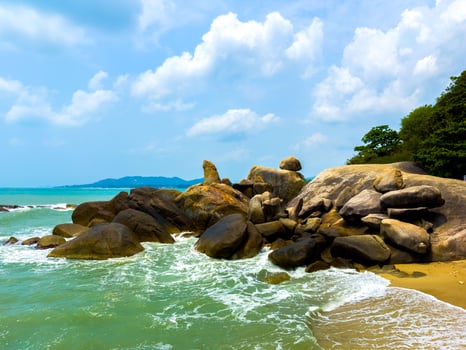 Wide-angle coastal landscape featuring the iconic Hin Ta and Hin Yai rock formations on Koh Samui, Thailand. The photo captures the calm turquoise water and white surf against the golden sand