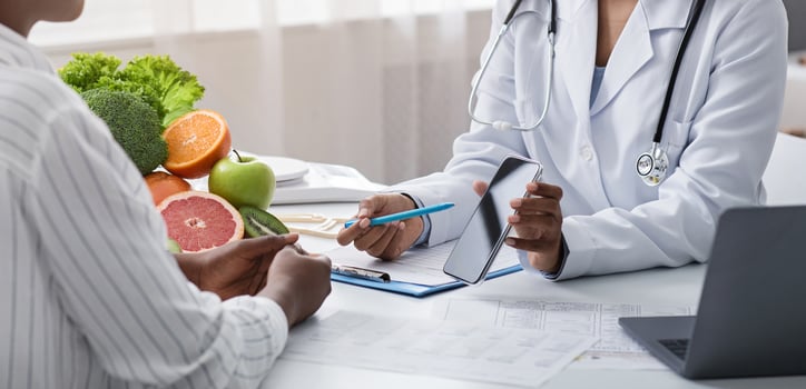 Mobile application for dieting. Nutritionist black woman showing patient blank smartphone screen, clinic interior, panorama photo