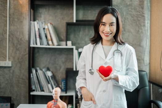 Female doctor in white coat holding heart model, standing in a modern medical office with books and anatomical model. photo