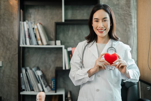 A cheerful female doctor in a white coat holds a heart model, standing in a contemporary office with a bookshelf. photo