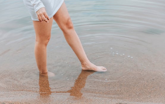 womans barefoot legs on wet sandy beach with feet in shallow water at lake shore wearing short white dress concept of wellness travel summer retreat fashion lifestyle photo