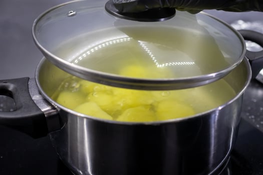 A black-gloved hand lifts the glass lid from a stainless steel pot of boiling potatoes, illustrating healthy home cooking and everyday meal preparation photo