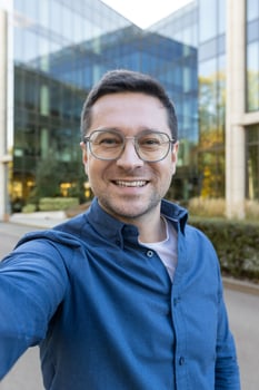 A man wearing glasses takes a selfie in front of a modern glass building, smiling towards the camera. photo