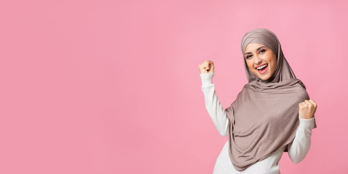 Great Bonus. Joyful arabic woman in headscarf celebrating success, raising hands and shaking fists over pink background, panorama with empty space photo