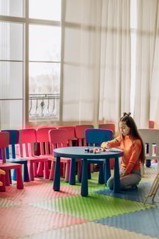 Girl painting alone at colorful table in creative classroom full of bright chairs and natural light showing peaceful artistic focus solitude and emotional self expression in early learning environment photo
