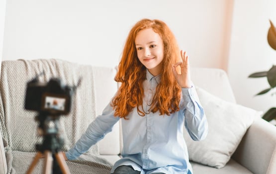 Blogging, Technology, Videoblog, Mass Media And People Concept. Happy smiling female teenage vlogger recording at home using camera on tripod and showing ok hand sign gesture, sitting on sofa photo