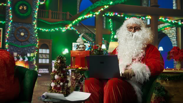 Smiling man dressed as Santa Claus looking at letter received from kids on laptop. Cheerful person wearing Santa costume reads wish lists written by children for Christmas on notebook device, camera A photo