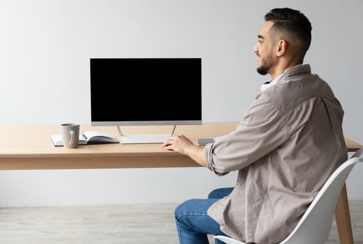 Rear back view of happy Middle Eastern man looking at blank empty computer monitor sitting at desk at workplace, watching webinar or having online web call, browsing internet, free copy space photo