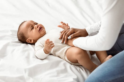 Portrait of irritated African American small infant crying and whining, suffering from colic or stomach pain, lying on back on white bedsheets, young mother doing belly massage for relief photo