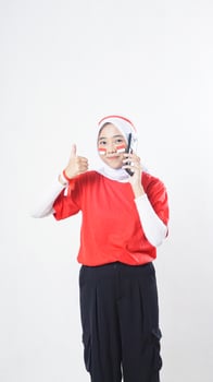 Indonesia independence day celebration with smiling Asian girl wearing red and white Indonesia flag color attribute talking on phone call while raising her thumb up isolated on white background photo