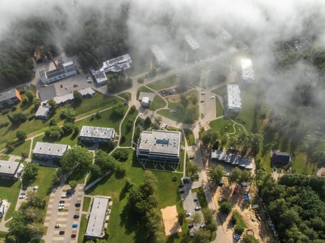 Cluster of modern buildings surrounded by trees and open green spaces, with pathways, parking lots, and light fog partially obscuring the scene. photo