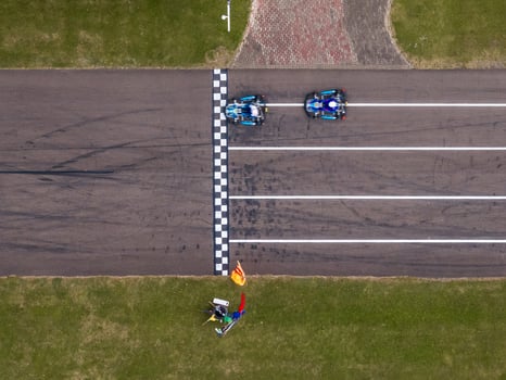 Two go karts cross a checkered finish line on a paved track, with a race official holding flags nearby. Tire marks and a brick walkway are visible. photo