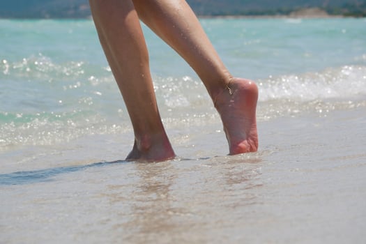 Close-up of female legs walking barefoot along the beach with sea waves. Wellness, travel and lifestyle concept. photo