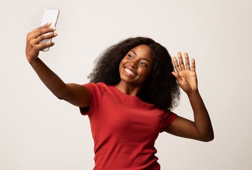 Happy Young Black Woman With Smartphone Making Call Or Taking Selfie, Portrait Of Cheerful African American Female Waving Hand At Mobile Phone Camera While Standing Over White Studio Background photo