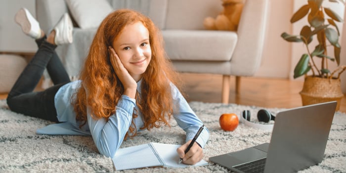 Studying At Home. Cheerful young female teenager lying on floor carpet with computer and taking notes in her notebook. Schoolgirl watching online course, learning foreign language looking at camera photo