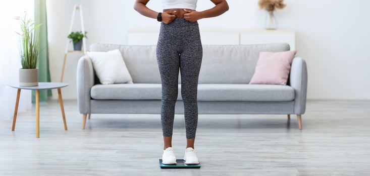 Weight Gain. Sad African American Lady Gaining Kilograms Touching Belly Standing On Scales At Home, Looking At Camera. Weight-Loss Problem, Visceral Fat And Slimming Issue Concept. Full Length photo