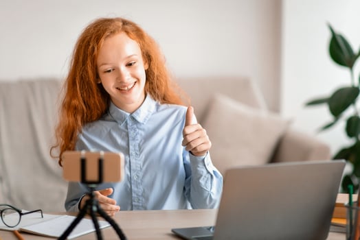Technology And Blogging. Positive young red-haired teen influencer girl shooting lifestyle vlog or review, talking to mobile camera on tripod, sitting at desk, showing thumbs up gesture, recommending photo