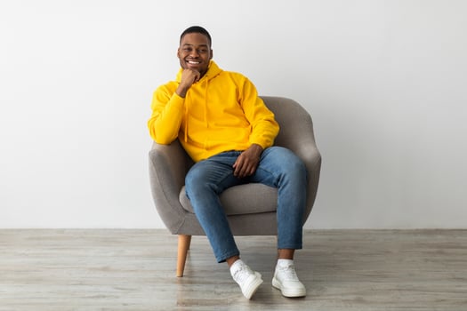Successful Black Millennial Guy Smiling To Camera Touching Chin Sitting In Armchair Near Gray Wall Indoors, Wearing Yellow Hoodie. African American Man Relaxing Posing At Home. Full Length photo