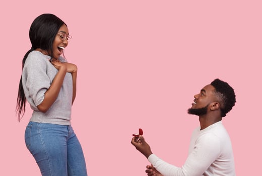 Black man in love making proposal to his girlfriend, staying on knee with ring, pink background, copy space photo