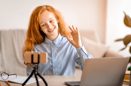Recording Review. Cheerful red-haired teenage girl influencer filming her blog talking to camera of smartphone on tripod, sitting at table with laptop. Smiling teen showing ok sign gesture photo