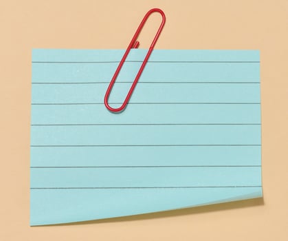 Blue sticky note with a red paper clip attached to it, placed on a beige background. Top view photo