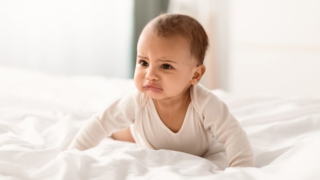 Portrait of crying African American infant in bodysuit whining, crawling on white bedsheets in bedroom at home indoors. Closeup of irritated baby feeling upset, banner, blurred background photo