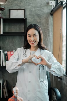 Cheerful female doctor in a white coat making a heart gesture, standing in a modern medical office. photo