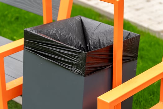 Trash bin with black liner and bright orange frame sits near a park bench, emphasizing urban public space cleanliness in sunny weather photo