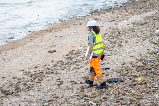 Environmental Scientist Inspecting Polluted Coastline, Worker in Safety Gear Walking on Rocky Beach with Debris, Female Engineer Surveying Coastal Area for Environmental Damage photo