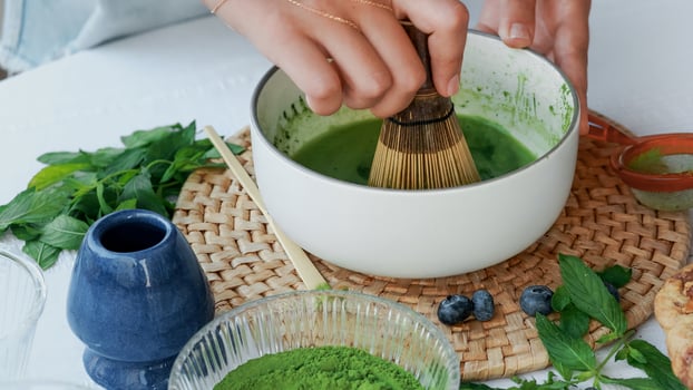 Homemade matcha ceremony. Green tea powder being whisked with water in a ceramic bowl using a bamboo whisk, surrounded by cookies, mint, and berries. Clean and mindful lifestyle concept photo