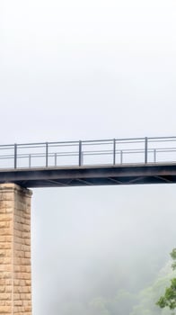 A stoic bridge emerges from a veil of mist, its robust structure and elegant railings piercing the ethereal fog, connecting two unseen worlds in a serene, atmospheric landscape. photo