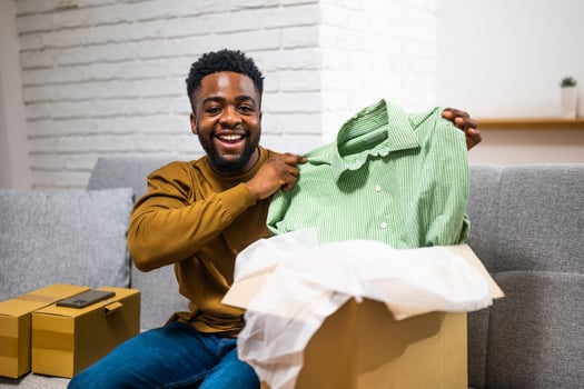 Young African American man happily unpacking clothes from a cardboard box delivered to his home, showcasing the convenience of online shopping. photo