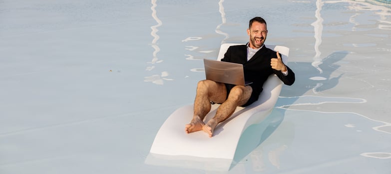 Man in blazer, swim trunks, and glasses sitting by the pool with a laptop, showing a thumbs-up gesture. Concept confidence in remote work and positive mindset photo