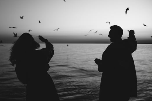 Silhouette of two people interacting with seagulls by ocean at dusk, with birds flying above. Black and white, soft light. Concept of freedom, nature, and joyful coastal moment. photo